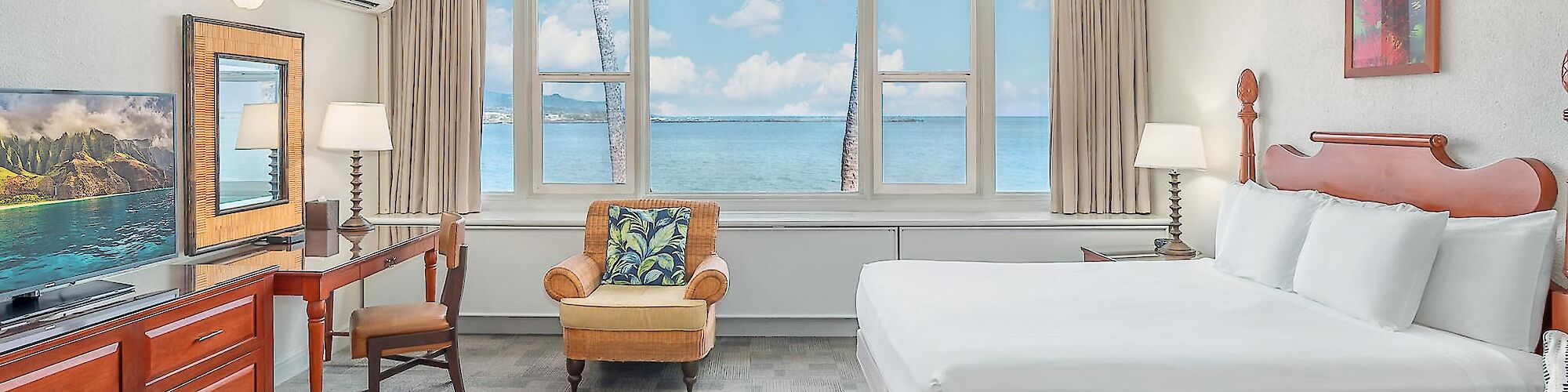 A spacious hotel room with a large bed, armchair, desk, TV, ceiling fan, and ocean view through the window, decorated with paintings.