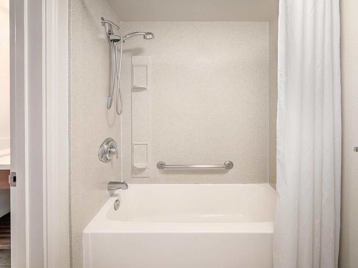 A clean, white bathtub with a showerhead, curtain, and grab bar in a bathroom. There are also built-in shelves for toiletries.