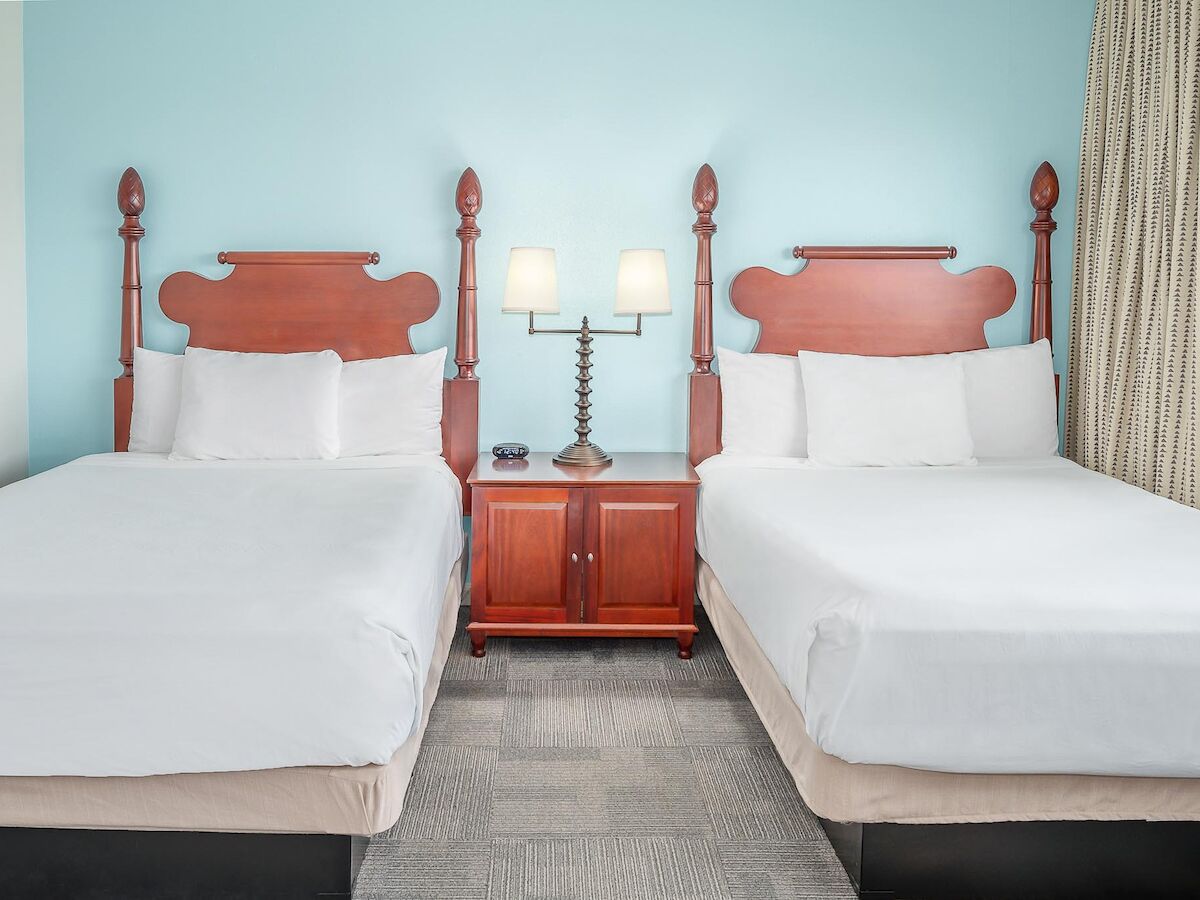 The image shows a hotel room with two neatly made beds, a central nightstand, and a lamp with two bulbs, all set against a light blue wall.