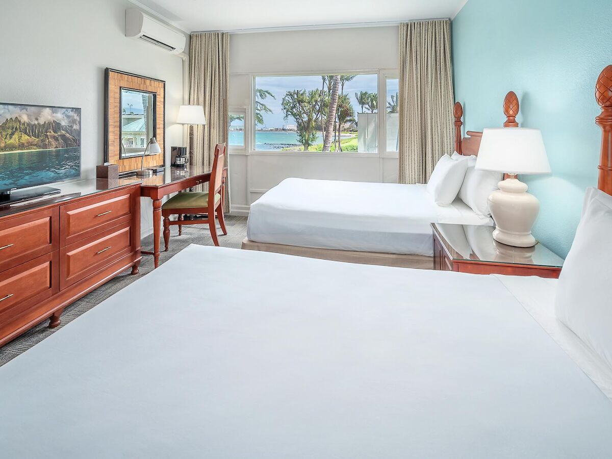 The image depicts a bright hotel room with two beds, a TV, a dresser, a desk with a chair, and a window with a view of the ocean and palm trees.