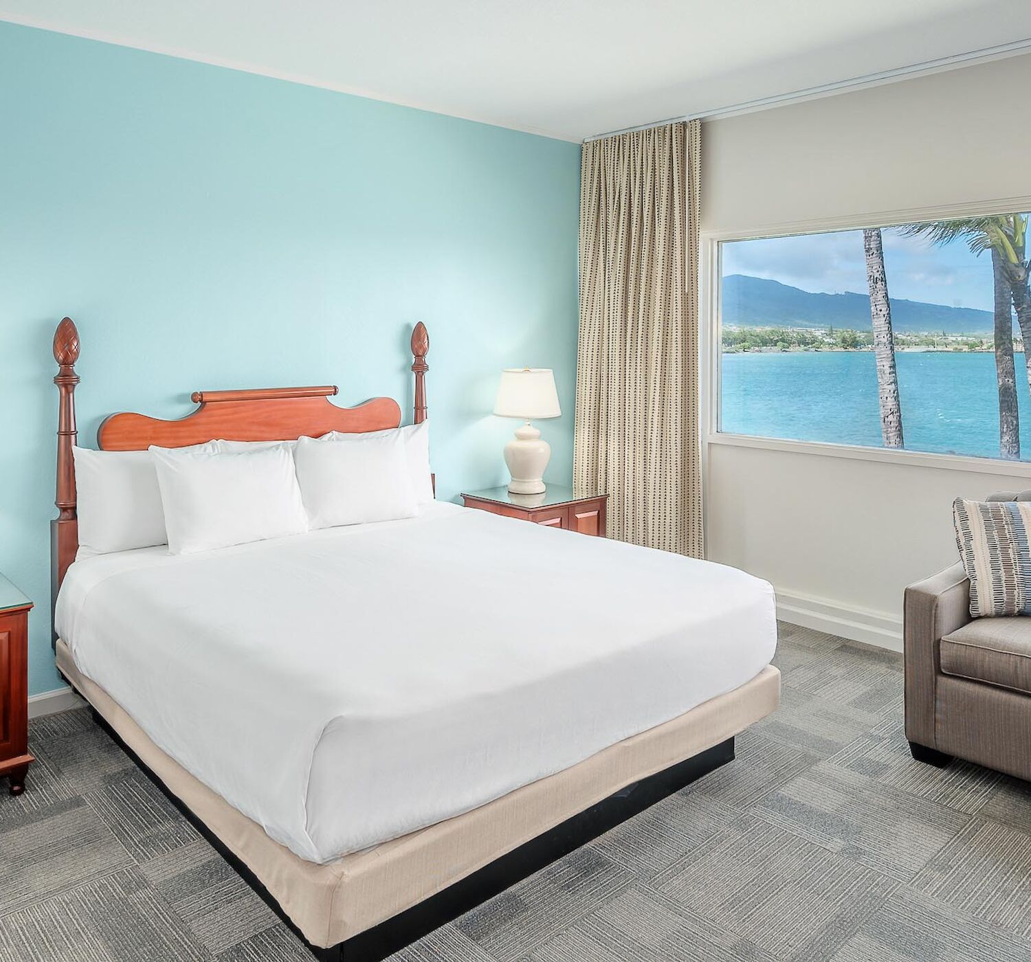 A bright, airy hotel room with a large bed, two nightstands with lamps, a sofa, and a window showcasing an ocean view with palm trees and mountains.