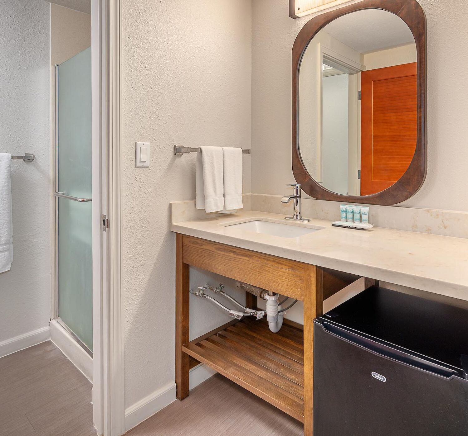 A bathroom with a vanity, mirror, sink, towels on a rack, small fridge, under-sink storage, and a shower separated by a glass door.