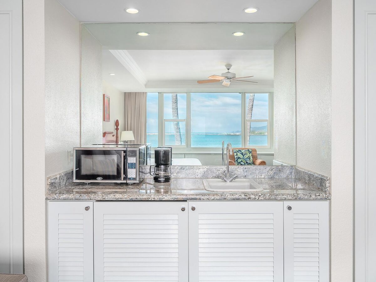 A kitchenette features a microwave, coffee maker, sink, and cabinets, with a large mirror reflecting a room with a view of the ocean and palm trees.