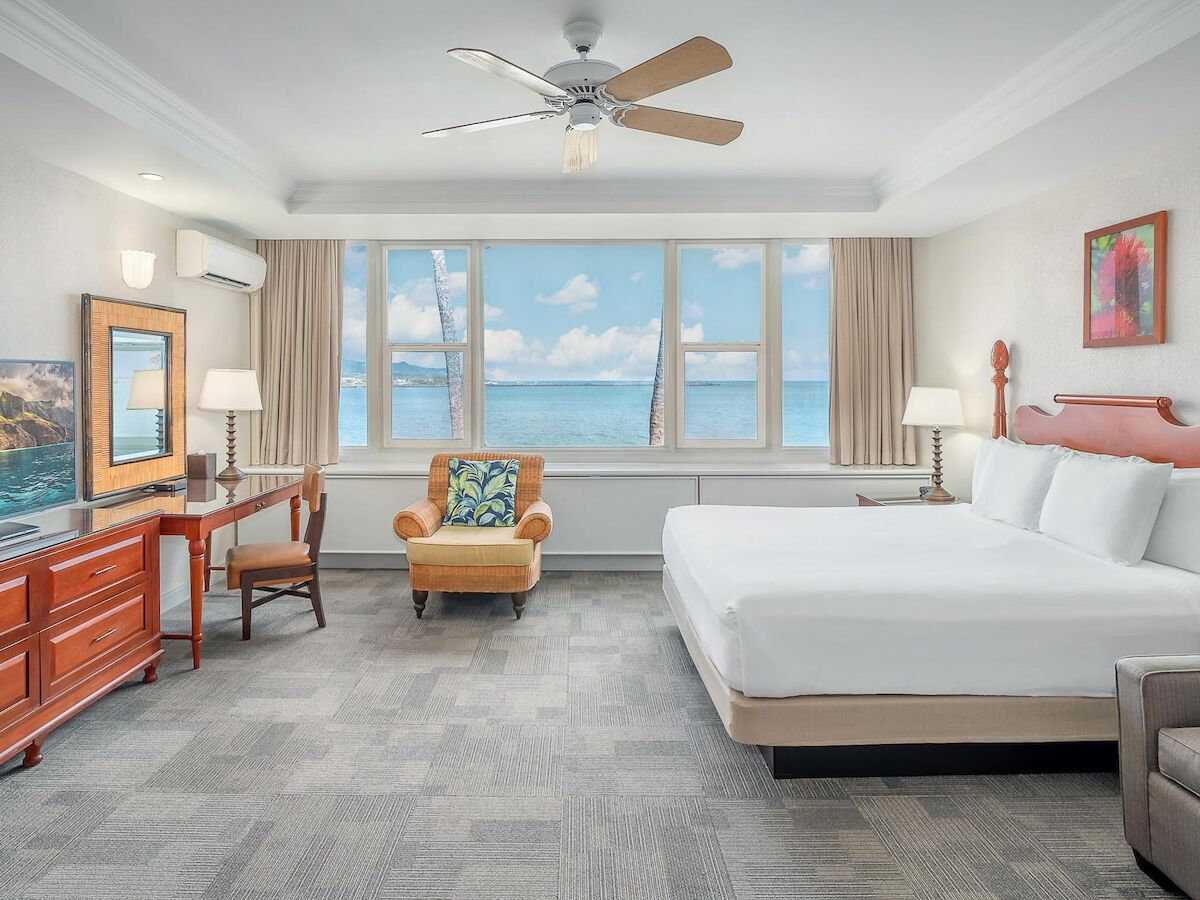 This image shows a bright, spacious hotel room with a large bed, TV, desk, armchair, and ocean view through the windows, ending the sentence.