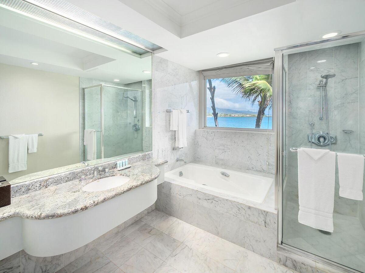 A modern bathroom features a large mirror, sink, bathtub, glass shower, and a window with an ocean view, all adorned with clean, white decor.