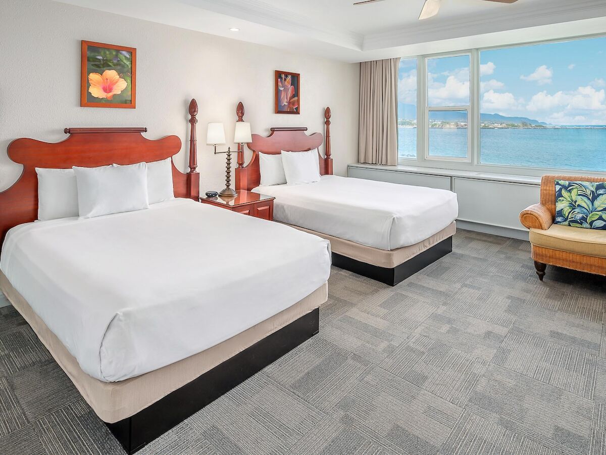 The image features a hotel room with two double beds, framed artwork, a nightstand, a chair, and a large window offering an ocean view.