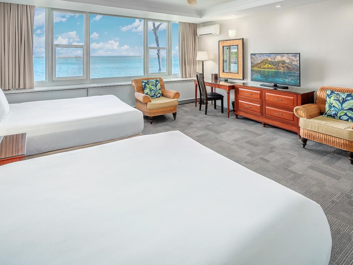A hotel room with two beds, a TV, two chairs, a desk, and large windows offering a scenic ocean view, creating a bright and inviting atmosphere.
