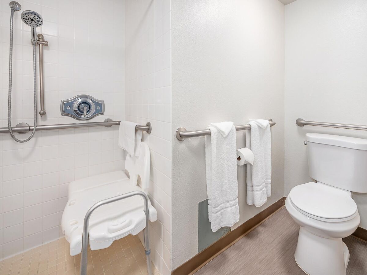 This image shows a bathroom with a toilet, a shower area with a bench, and grab bars, designed for accessibility, with white towels hanging on the bars.