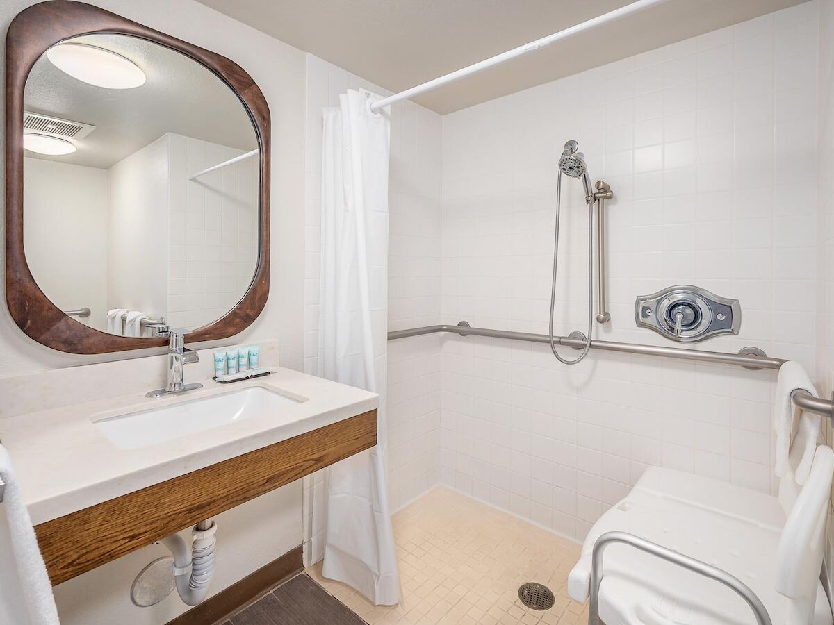 This image shows an accessible bathroom with a sink, mirror, shower with grab bars, shower seat, white tiles, and toiletries.