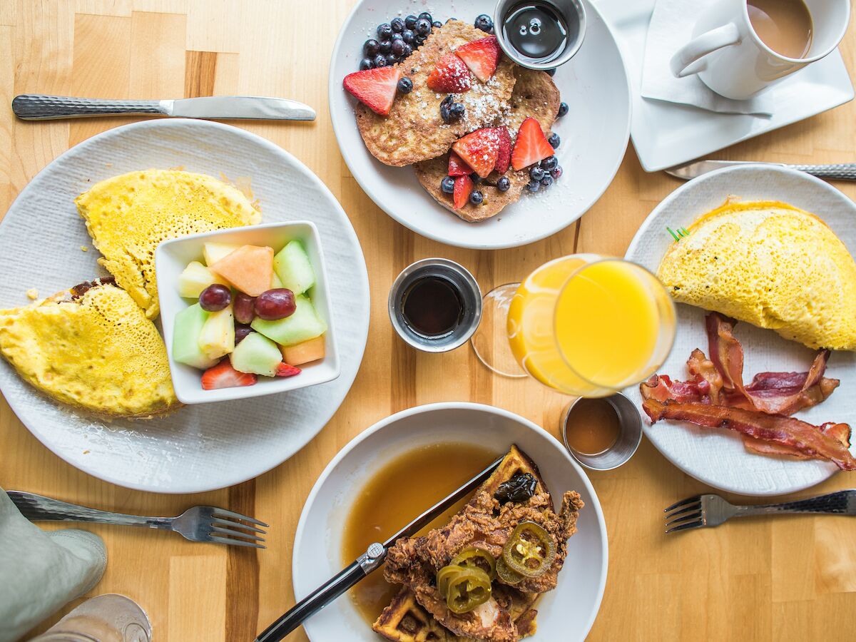 The image shows a breakfast spread with omelettes, waffles, fruit salad, pancakes with berries, bacon, orange juice, coffee, and syrup.
