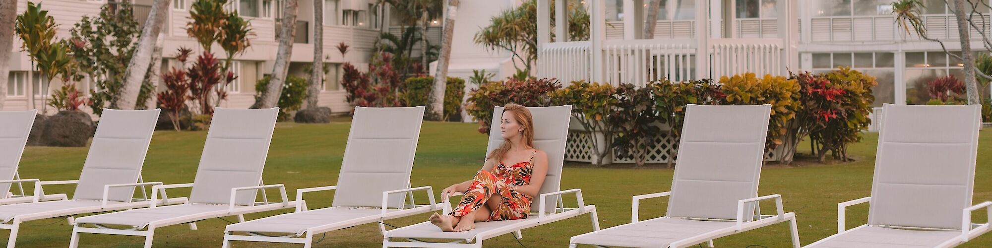 A woman in a floral dress is lounging on a white chair, surrounded by empty chairs in a garden with a gazebo and a building in the background.