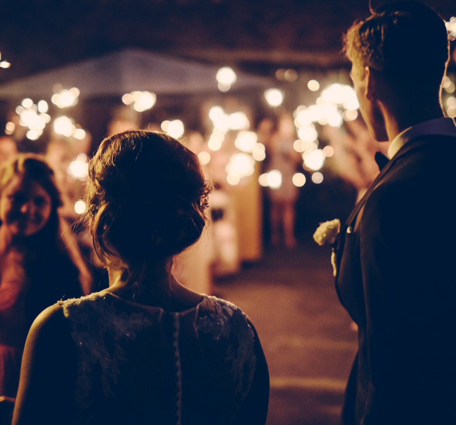 A couple in formal attire is facing a crowd at night. The crowd holds sparklers, creating a festive, warm atmosphere, possibly at a celebration.