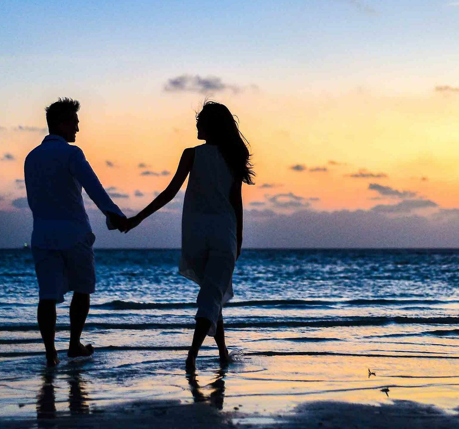 A couple holding hands walks along a beach at sunset, with calm waves and a colorful sky in the background.