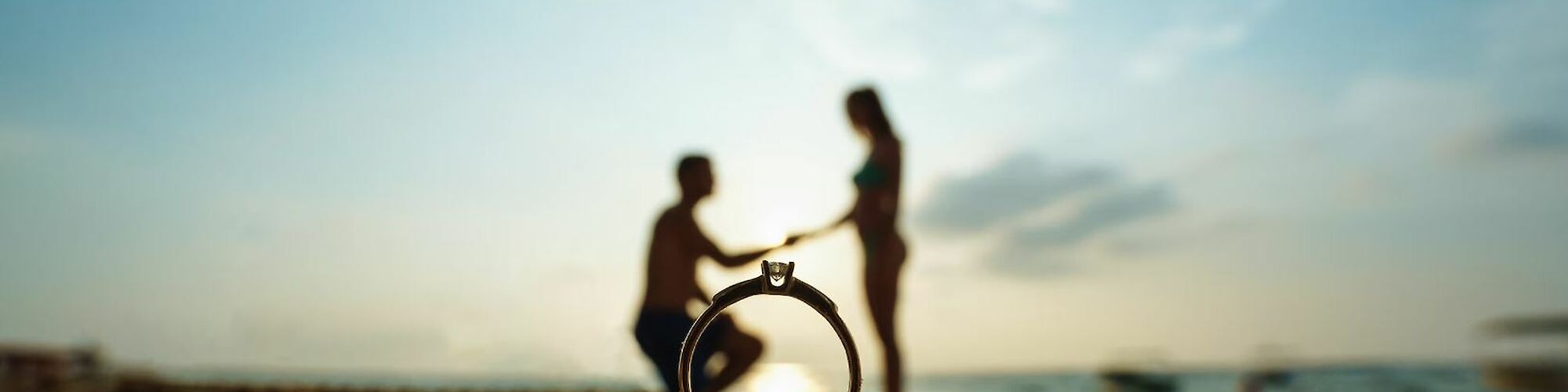 A ring is in focus on sandy beach, with a silhouetted couple in the background, one kneeling, likely proposing, during a beach sunset.
