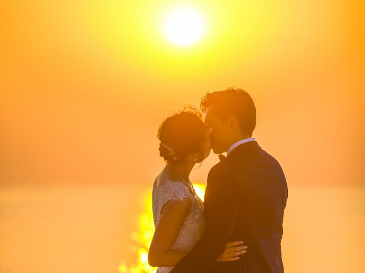 A couple dressed in wedding attire shares a romantic moment on the beach during a stunning sunset with the sun reflecting on the water's surface.