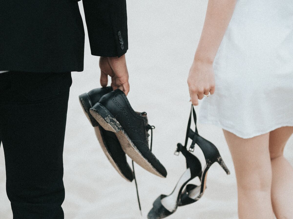 A couple in formal attire walks barefoot on a sandy beach, each holding their shoes in hand.
