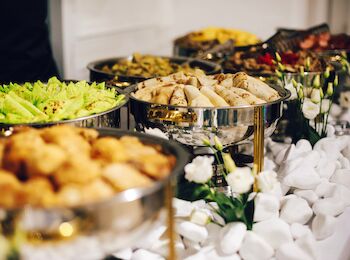 The image shows a buffet table with various plated dishes including spring rolls, salads, and other appetizers, adorned with flowers.