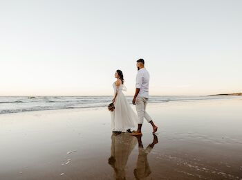 A couple is walking barefoot on a beach, with the woman in a white dress and the man in light-colored pants and a white shirt, holding hands.