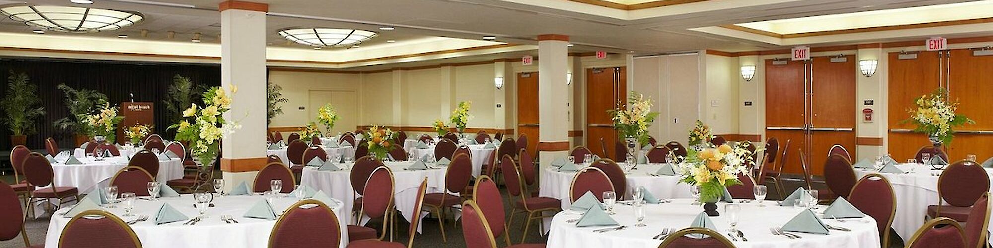 A banquet hall with round tables set for an event, featuring white tablecloths, arranged chairs, and floral centerpieces, under soft lighting.
