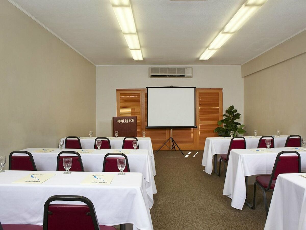 A small conference room set up with white tablecloths, chairs, a projector screen, a podium, and a potted plant in the corner.
