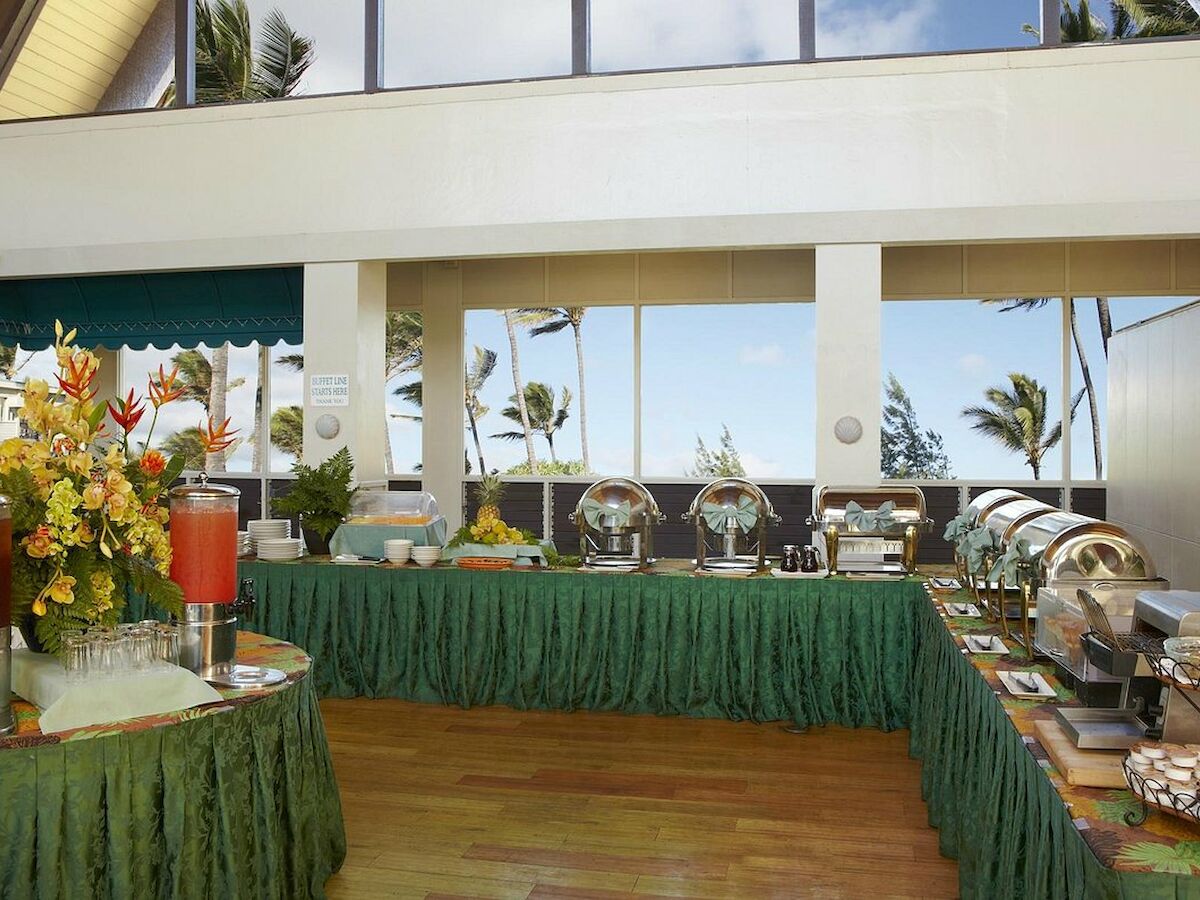 A buffet setup with chafing dishes, beverages, and tropical decor is displayed indoors with a view of palm trees outside the windows.