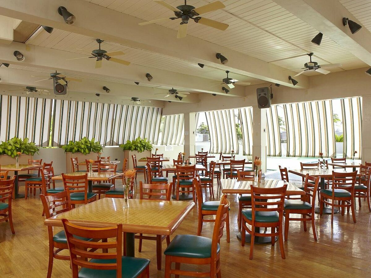 The image shows an empty indoor dining area with wooden tables and chairs, ceiling fans, and large windows bringing in natural light.