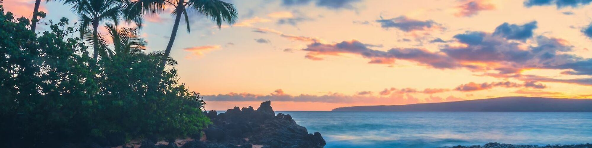 A tropical beach scene at sunset with palm trees, rocky shoreline, calm waves, and colorful sky.