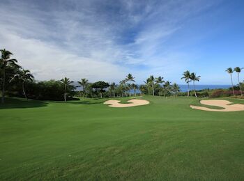 A scenic golf course featuring lush green fairways, sand bunkers, and palm trees under a partly cloudy sky near the ocean.