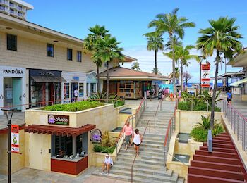 An outdoor shopping area with palm trees, shops, and people walking down stairs. Brands like Pandora are visible. Bright and sunny day.