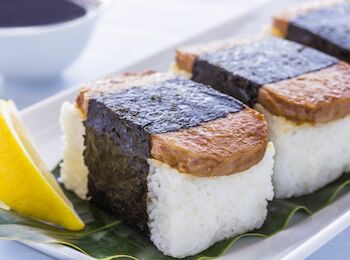 The image shows slices of Spam musubi, a Hawaiian dish, placed with a slice of lemon on a white plate. A bowl with dark sauce is in the background.