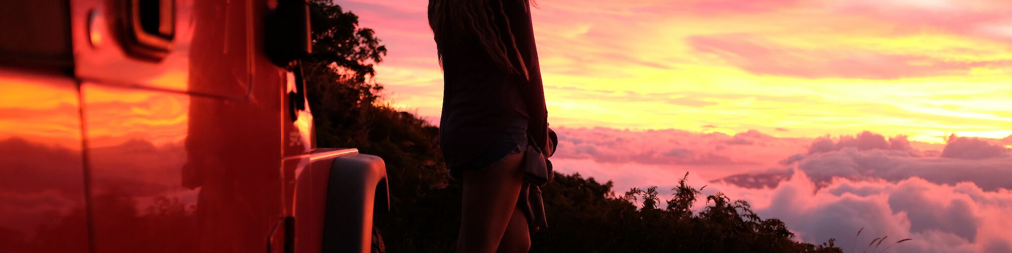 A person is standing near a vehicle, gazing at a vibrant sunset above the clouds, with a breathtaking view in the background.