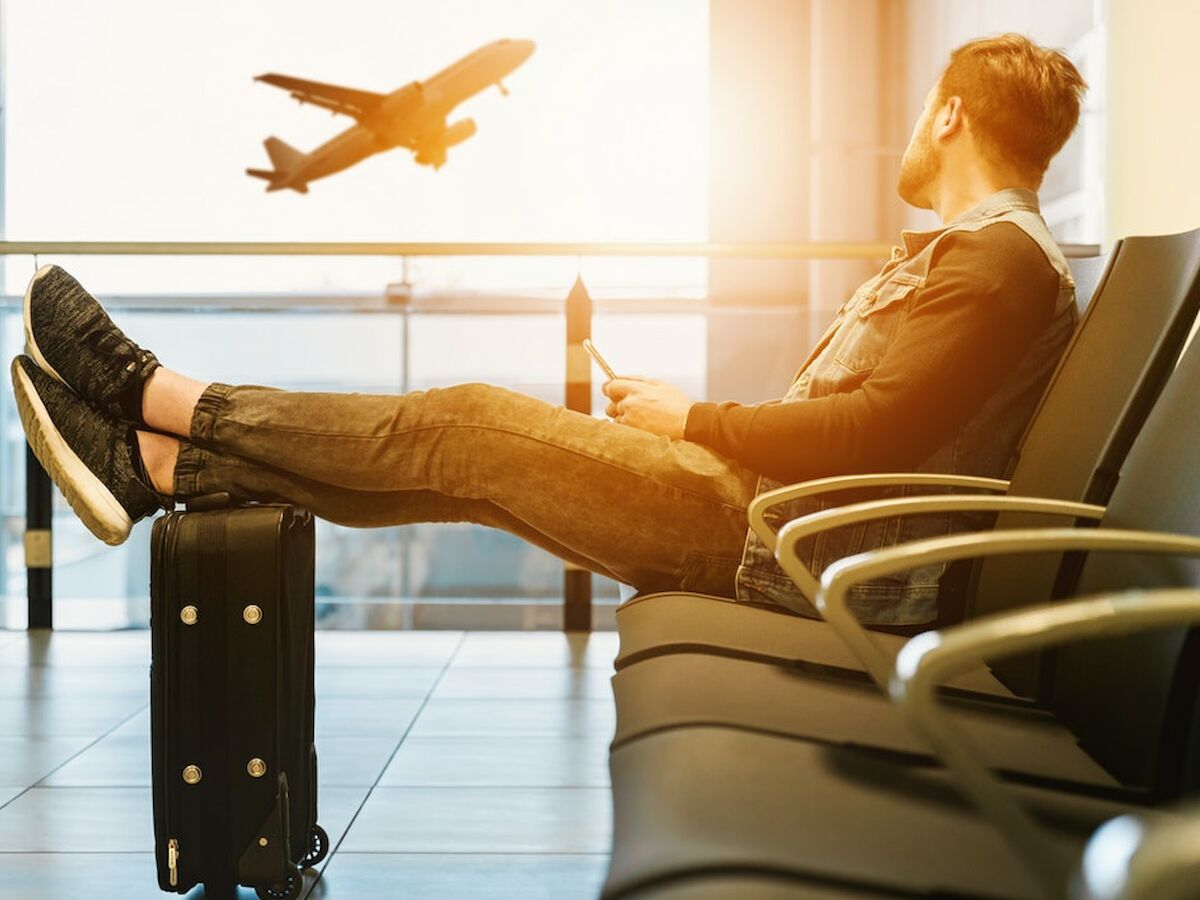 A person sits in an airport lounge with their feet on a suitcase, watching a plane take off through large windows.