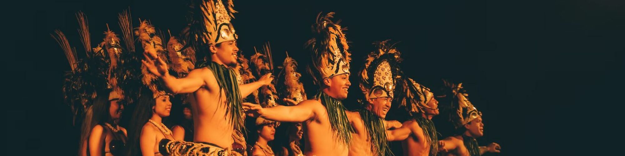 A group of people in traditional attire, performing a cultural dance with feathered headdresses and grass skirts, set against a dark background.