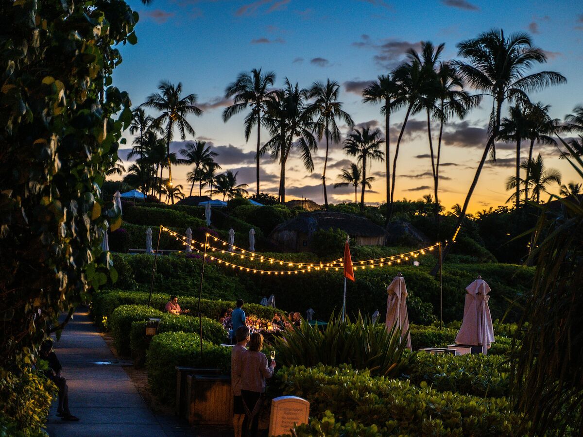The image shows a tropical outdoor setting at dusk with string lights, palm trees, and people dining among lush greenery and a vivid sunset sky.