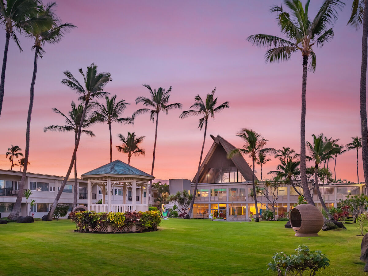 A sunset over a tropical resort with palm trees, a gazebo, and buildings. The sky is colorful with shades of pink and purple, enhancing the scenery.