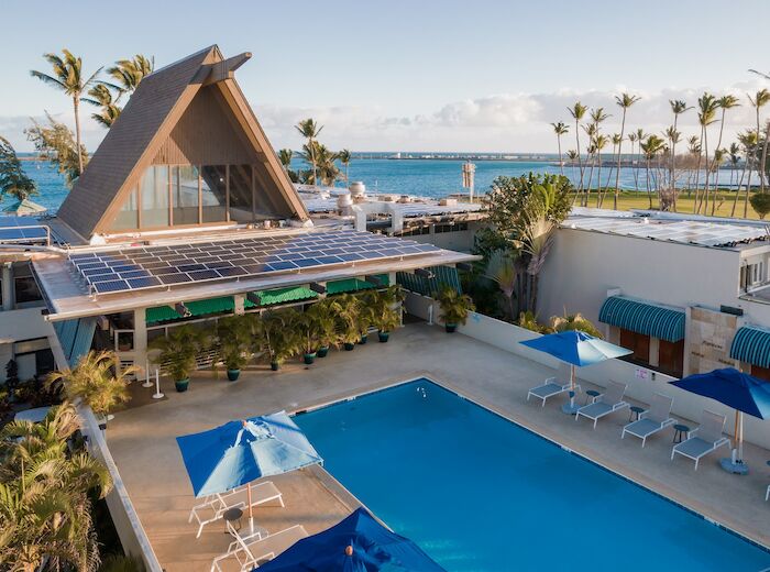 An outdoor swimming pool with lounge chairs and umbrellas, adjacent to a modern building with an A-frame roof and solar panels, near the ocean.
