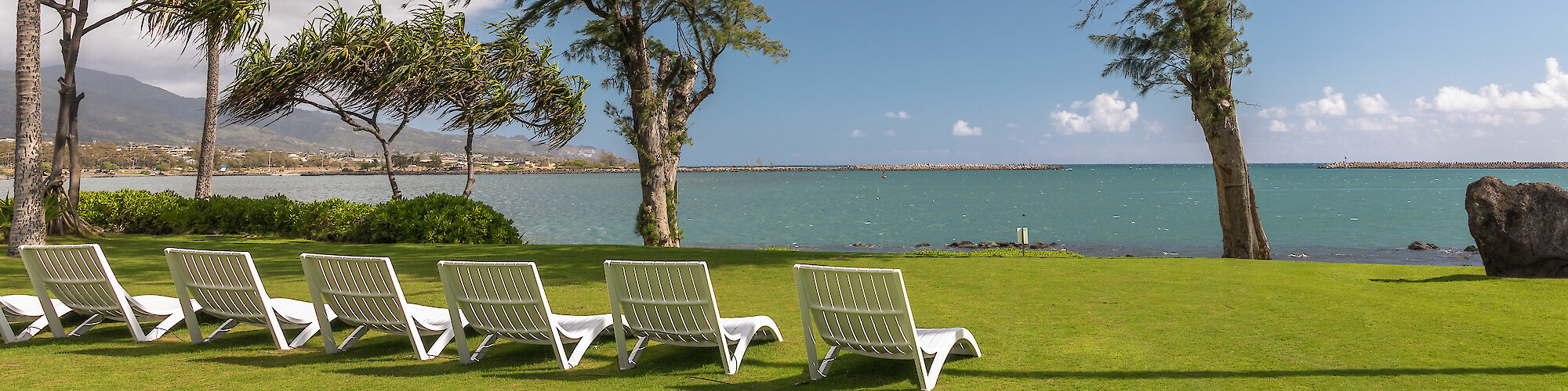 The image shows a row of white lounge chairs on a green lawn, overlooking a scenic ocean view with trees and distant mountains.