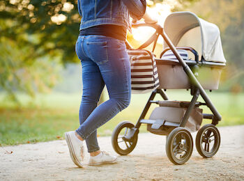 A person in jeans and sneakers is pushing a baby stroller on a path in a park with sunlight streaming through the trees in the background.
