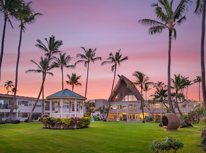 A beautifully landscaped resort with palm trees, a gazebo, a modern building, and a sunset sky in the background.