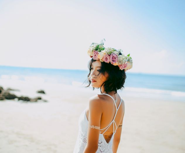 A person in a white dress with a flower crown stands on a beach, looking towards the ocean under a clear blue sky, creating a serene scene.