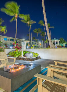 This image shows an outdoor seating area with Adirondack chairs around a fire pit, tropical plants, and palm trees at night.