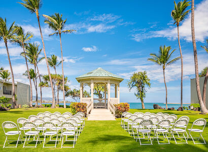 The image shows a wedding setup with white chairs facing a gazebo on a lush lawn, surrounded by palm trees, overlooking the ocean under a blue sky.
