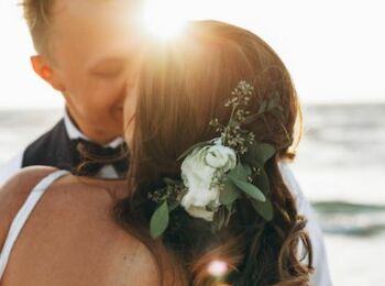 A couple is embracing on a beach at sunset, with flowers in the woman's hair and the sun shining above them.