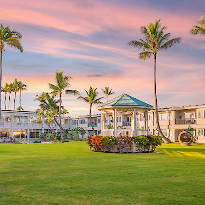 The image shows a beautifully landscaped resort with a large lawn, palm trees, a central gazebo, and buildings under a colorful sky.