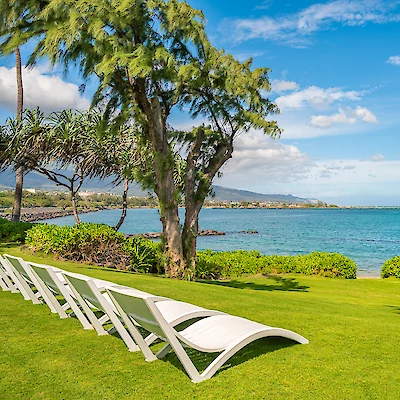 The image shows a beautiful coastal landscape with several white lounge chairs on lush green grass, overlooking the serene blue ocean under a clear sky.