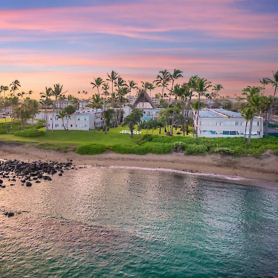 A scenic coastal view with a beach, palm trees, and buildings under a colorful sunset sky. There are rocks in the shallow, clear water near the shore.
